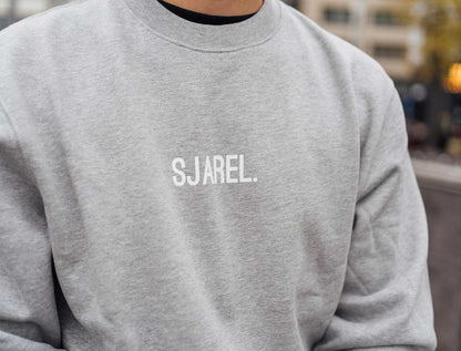 Close-up van een grijze sweater met opdruk SJAREL.
