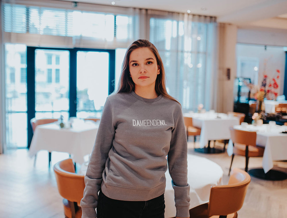 Vrouw met een grijze trui met opdruk DAMEENDENI.