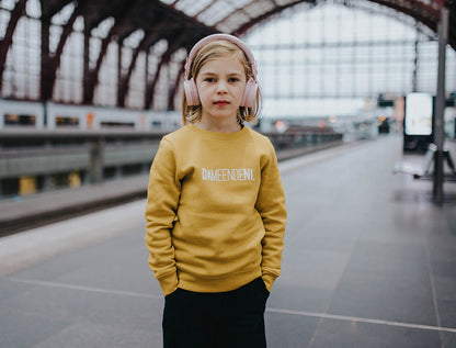 Meisje met okerkleurige trui met opdruk DAMEENDENI.