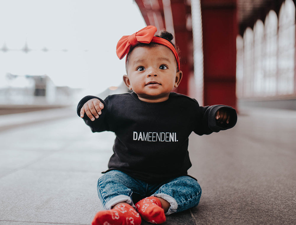 Baby met zwarte trui met opdruk DAMEENDENI.